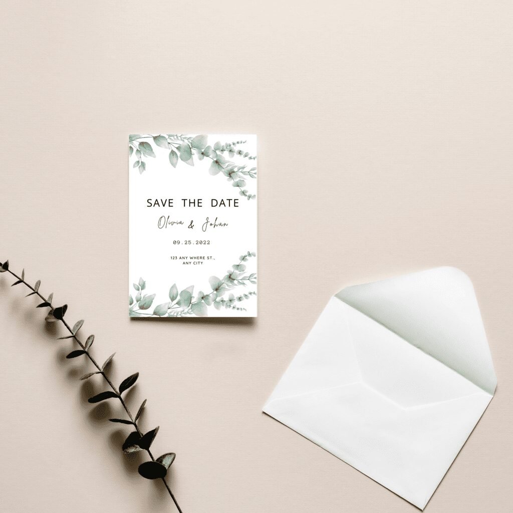 A Save the Date card with green leafy designs lies on a beige surface next to a white open envelope and an eucalyptus branch. The elegant fonts and botanical wedding clipart reflect the latest wedding trends.