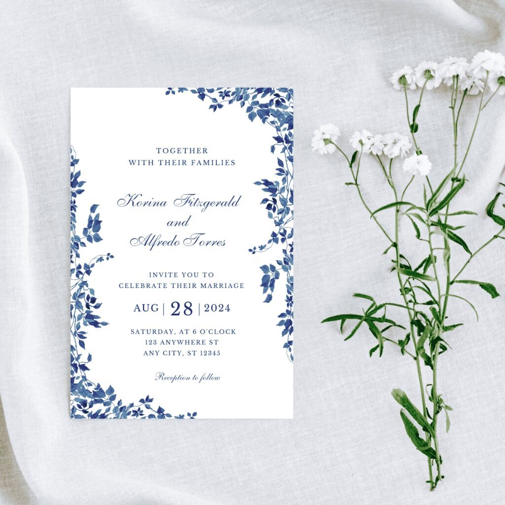 A wedding invitation with blue floral borders, inspired by the latest wedding trends, sits on light fabric next to small white flowers. The invitation is for Korina Fitzgerald and Alfredo Torres, with the event on 28 August 2024 at 6 o’clock.