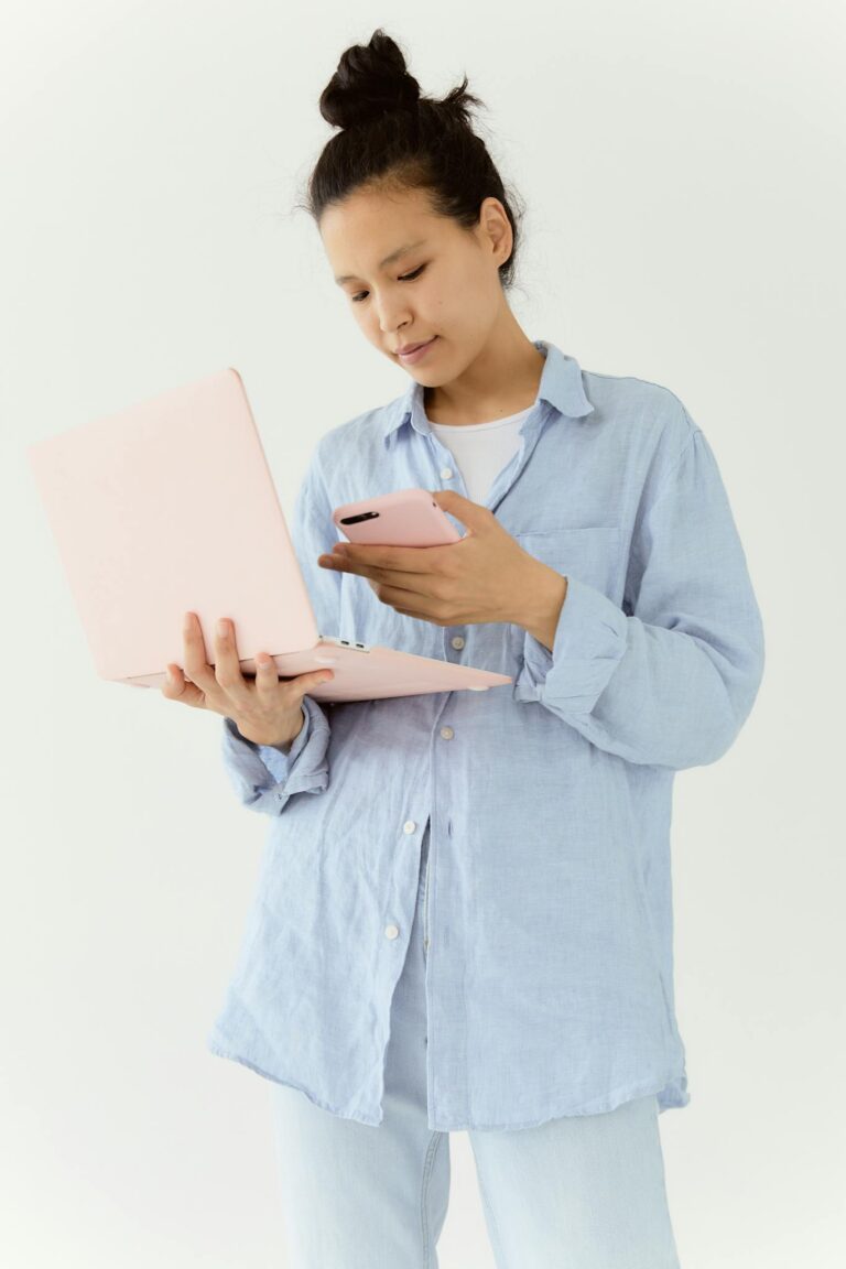 Asian woman in blue shirt multitasking with a laptop and smartphone indoors.