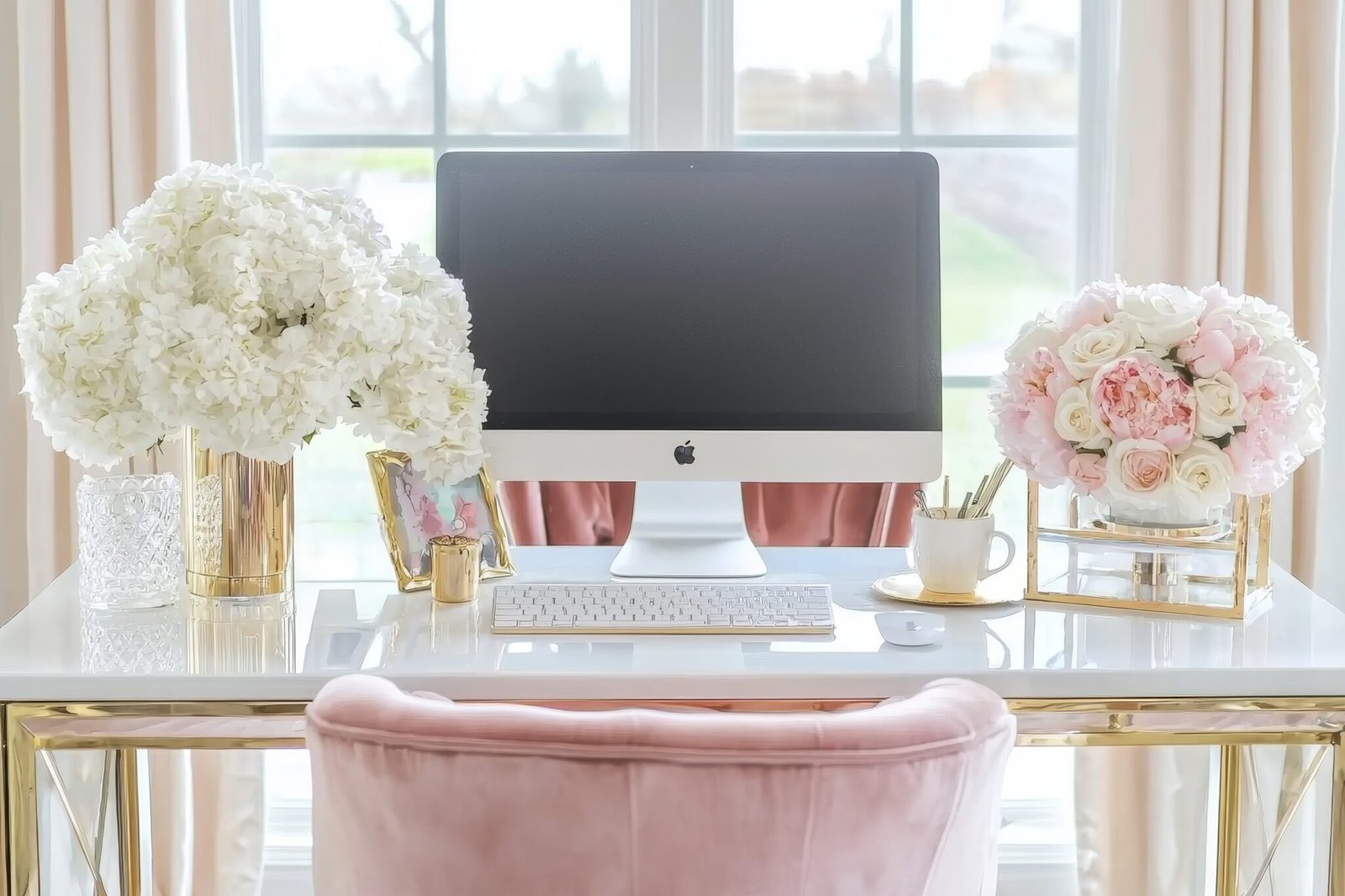 A stylish home office desk with an iMac, white keyboard, mouse, white and pink floral arrangements in gold vases, stationery, and a framed photo, set in front of large windows with soft pink curtains.