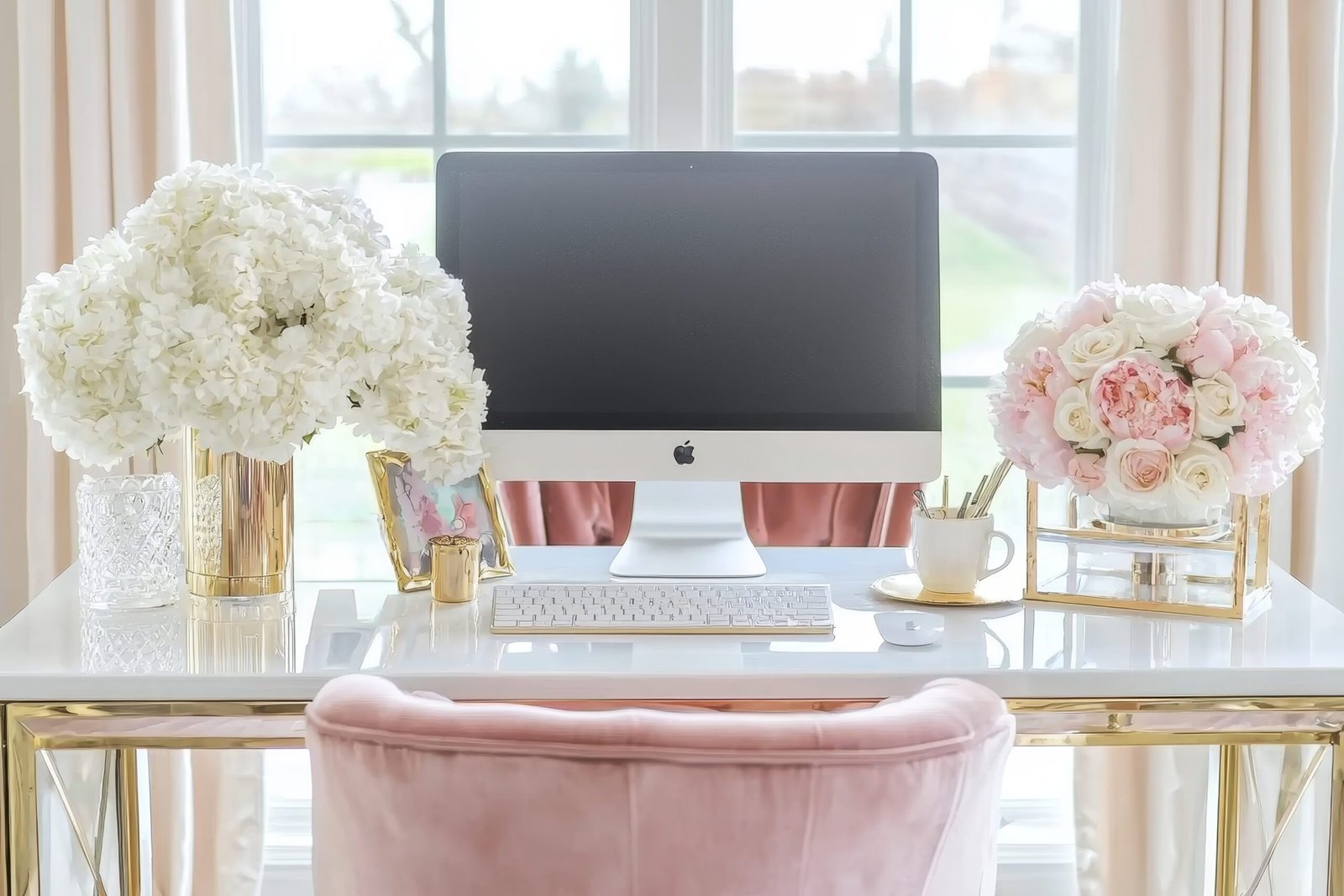 A stylish home office desk with an iMac, white keyboard, mouse, white and pink floral arrangements in gold vases, stationery, and a framed photo, set in front of large windows with soft pink curtains.